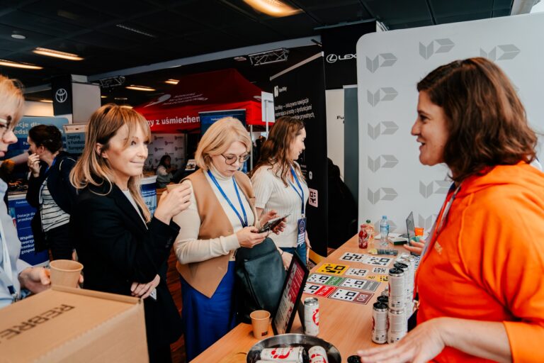 Przedstawicielka Urzędu Patentowego Rzeczypospolitej Polskiej w pomarańczowej bluzie rozmawia z uczestniczkami 15. Forum Rozwoju Mazowsza przy stoisku informacyjnym. Na stole leżą materiały promocyjne i napoje.