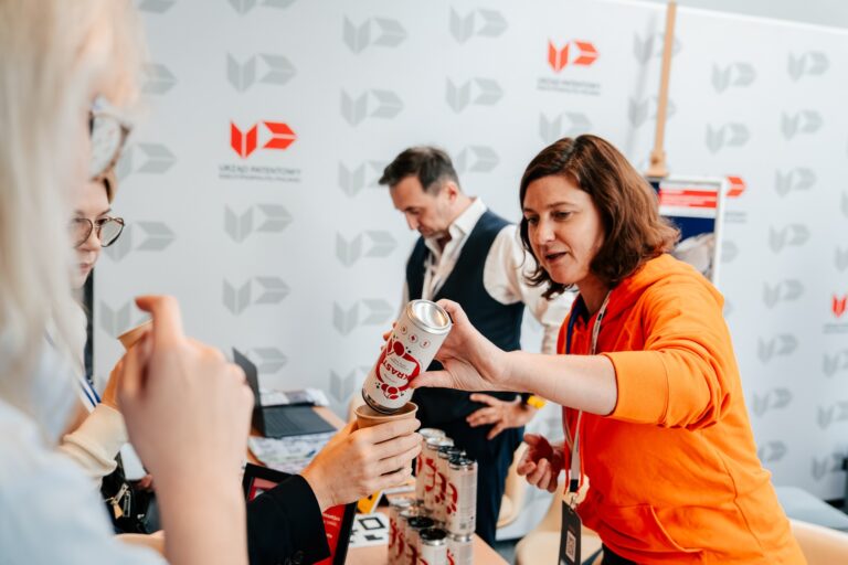 Przedstawicielka Urzędu Patentowego Rzeczypospolitej Polskiej w pomarańczowej bluzie podaje napój uczestniczce wydarzenia podczas 15. Forum Rozwoju Mazowsza. W tle widać stoisko urzędu i inne osoby.