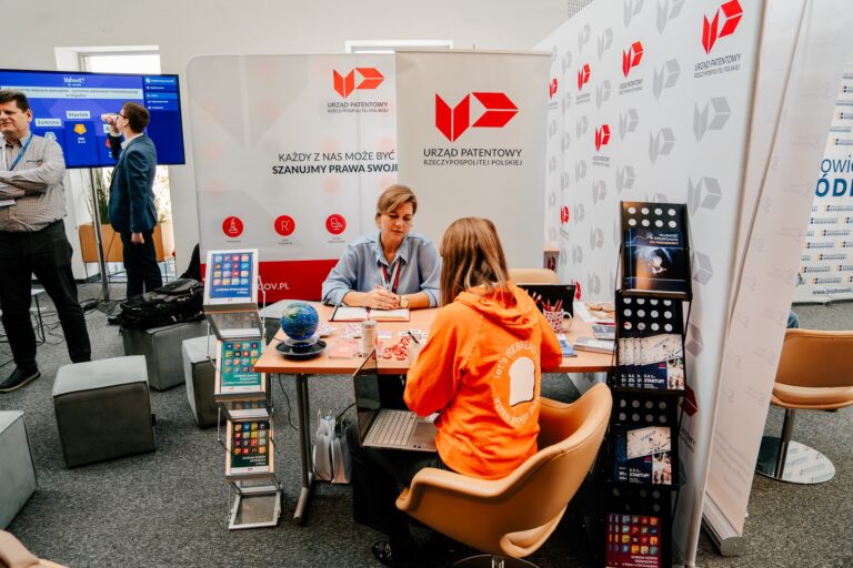 Stoisko Urzędu Patentowego Rzeczypospolitej Polskiej podczas 15. Forum Rozwoju Mazowsza. Przedstawicielka urzędu rozmawia z uczestniczką wydarzenia w pomarańczowej bluzie. W tle widoczne są materiały informacyjne i ekran prezentacyjny.