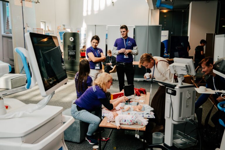Grupa osób w fioletowych koszulkach prowadzi pokaz medyczny z użyciem fantomów niemowląt. Uczestnicy obserwują demonstrację resuscytacji i badań z wykorzystaniem sprzętu medycznego. Scena odbywa się w jasnym pomieszczeniu podczas wydarzenia edukacyjnego.