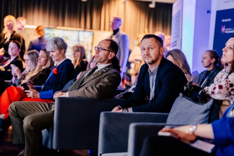 Uczestnicy 15. Forum Rozwoju Mazowsza siedzą na widowni i słuchają prezentacji. W pierwszym rzędzie widoczny jest mężczyzna w mundurze wojskowym oraz inni uczestnicy wydarzenia w skupieniu.