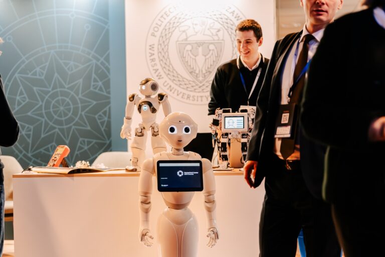 Robot humanoidalny prezentowany na stoisku Politechniki Warszawskiej podczas 15. Forum Rozwoju Mazowsza. W tle widać inne roboty, uczestników wydarzenia i logo uczelni.
