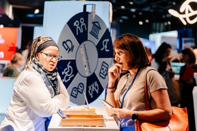 wie kobiety rozmawiają przy stoisku podczas 15. Forum Rozwoju Mazowsza. W tle widać koło z ikonami przedstawiającymi różne obszary tematyczne oraz innych uczestników wydarzenia.