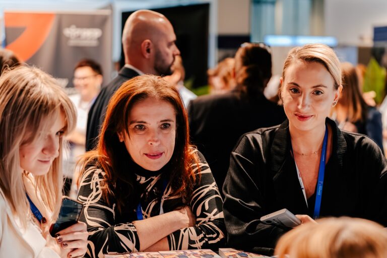 Trzy kobiety siedzą przy stole na wydarzeniu branżowym, rozmawiają i przeglądają materiały. W tle widoczni są inni uczestnicy spotkania w przestrzeni wystawienniczej.
