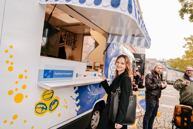 Kobieta stoi przy foodtrucku oznaczonym logotypami Funduszy Europejskich i programu regionalnego. Uśmiecha się do aparatu, trzymając kubek z napojem. W tle widoczni są inni uczestnicy wydarzenia i kolejne pojazdy gastronomiczne ustawione wzdłuż ulicy.