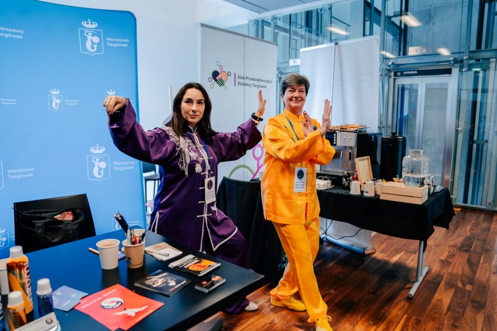 Dwie kobiety w kolorowych strojach do tai chi – jedna w fioletowym, druga w żółtym – pozują w dynamicznych pozycjach przed ścianką z napisem „Warszawa Targówek”. W tle widoczne stoisko Klubu Przedsiębiorczości Dzielnicy Targówek z ekspresem do kawy i materiałami promocyjnymi.