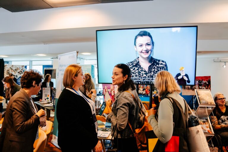 Uczestnicy wydarzenia rozmawiają przy stoisku informacyjnym. W tle duży ekran wyświetla uśmiechniętą kobietę w koszuli w panterkę oraz napisy promocyjne. Na stołach widoczne są broszury i materiały informacyjne, a w tle kilka innych stoisk targowych.