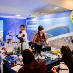 Stoisko Podkarpackiego Centrum Nauki Łukasiewicz podczas wydarzenia. Kilka osób uczestniczy w pokazach naukowych – na stole widać sprzęt laboratoryjny, probówki, pipety i rośliny. Prowadząca w fartuchu laboratoryjnym i druga osoba w kolorowej koszuli tłumaczą przebieg doświadczeń uczestnikom. W tle baner Fundusze Europejskie dla Podkarpacia 2021–2027.