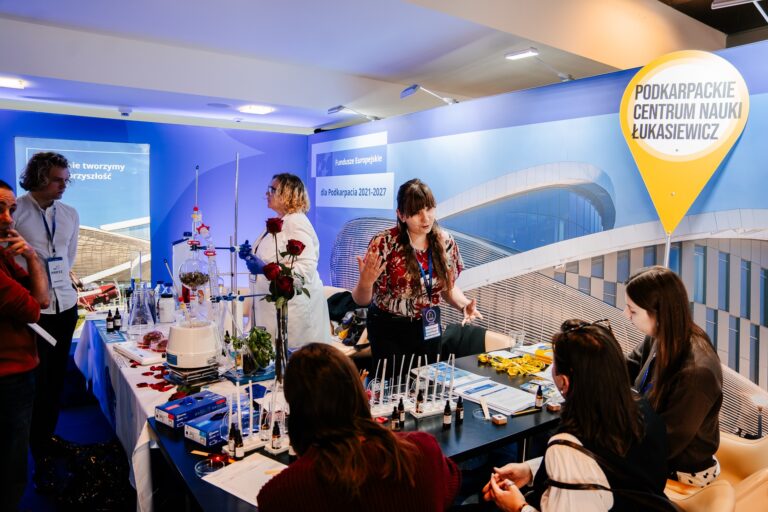 Stoisko Podkarpackiego Centrum Nauki Łukasiewicz podczas wydarzenia. Kilka osób uczestniczy w pokazach naukowych – na stole widać sprzęt laboratoryjny, probówki, pipety i rośliny. Prowadząca w fartuchu laboratoryjnym i druga osoba w kolorowej koszuli tłumaczą przebieg doświadczeń uczestnikom. W tle baner Fundusze Europejskie dla Podkarpacia 2021–2027.