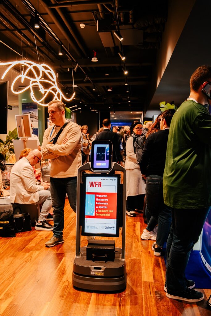 Mobilny robot informacyjny z ekranem dotykowym prezentuje komunikat o wsparciu finansowym z Funduszy Europejskich. Urządzenie stoi na korytarzu wydarzenia pełnym uczestników, w tle widać stoiska wystawowe i osoby rozmawiające.