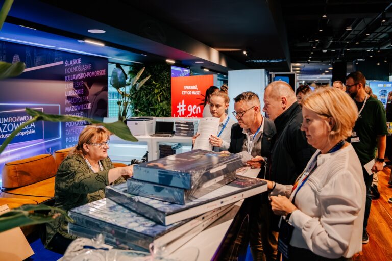 Kilka osób stoi przy ladzie informacyjnej podczas wydarzenia, odbierając materiały lub gadżety. Kobieta za ladą przekazuje zapakowane przedmioty, a uczestnicy przeglądają dokumenty. W tle widoczne są kolorowe stoiska i podświetlone napisy związane z Funduszami Europejskimi