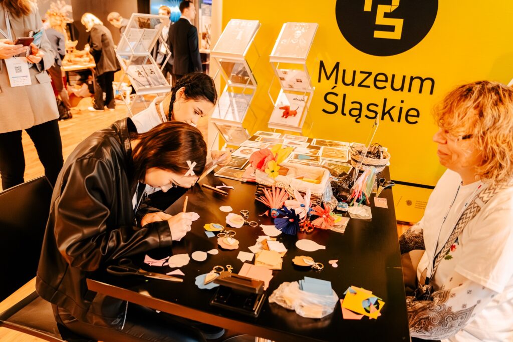 Kobieta siedzi przy stole i wycina kolorowe elementy z papieru podczas warsztatów artystycznych. Na stole widać przybory plastyczne oraz papierowe kwiaty, a w tle znajdują się stojaki z broszurami i uczestnicy wydarzenia.