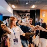 Młody mężczyzna w kapeluszu i młoda kobieta stoją wśród uczestników wydarzenia, trzymając na rękawicach ptaki drapieżne – sowę i sokoła. W tle widać innych uczestników konferencji oraz korytarz z oświetleniem sufitowym.