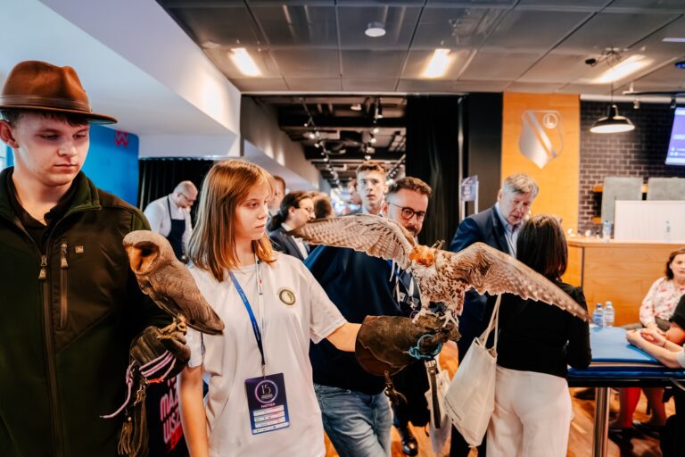 Młody mężczyzna w kapeluszu i młoda kobieta stoją wśród uczestników wydarzenia, trzymając na rękawicach ptaki drapieżne – sowę i sokoła. W tle widać innych uczestników konferencji oraz korytarz z oświetleniem sufitowym.