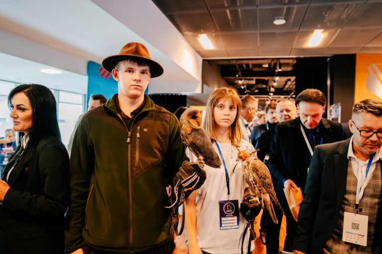 Młody mężczyzna w kapeluszu i młoda kobieta stoją wśród uczestników wydarzenia, trzymając na rękawicach ptaki drapieżne – sowę i sokoła. W tle widać innych uczestników konferencji oraz korytarz z oświetleniem sufitowym.