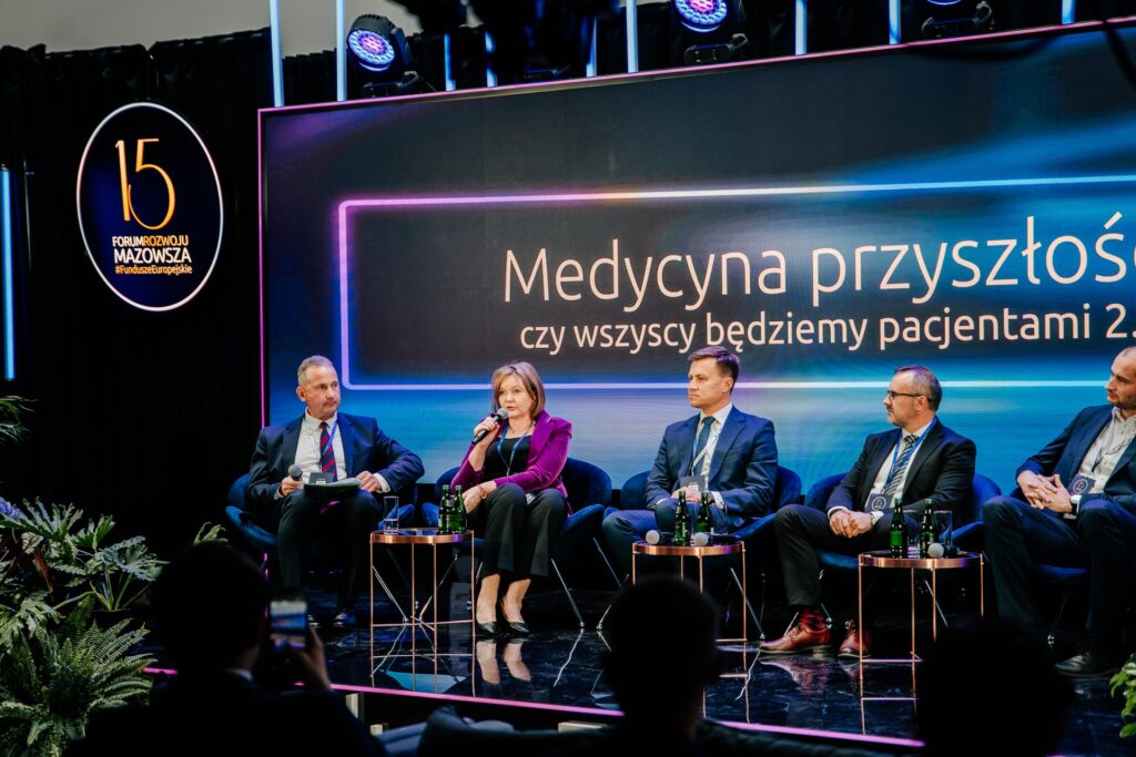 Uczestnicy debaty „Medycyna przyszłości: czy wszyscy będziemy pacjentami 2.0?” siedzą na scenie podczas 15. Forum Rozwoju Mazowsza. Kobieta w fioletowej marynarce mówi do mikrofonu, a obok niej siedzą mężczyźni w garniturach. W tle widoczny jest ekran z tytułem dyskusji i logo wydarzenia.
