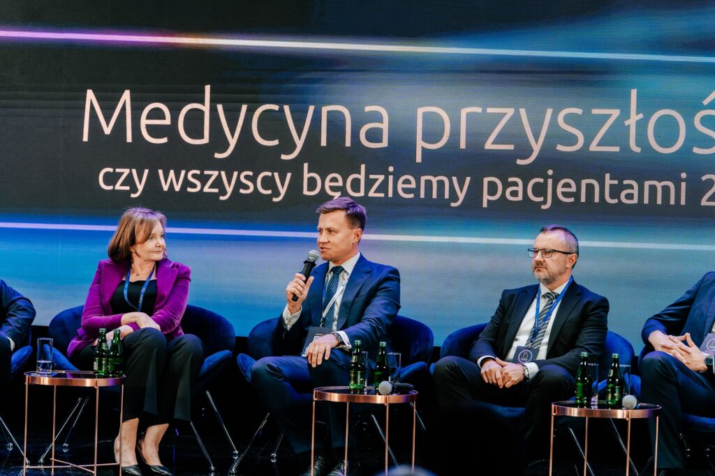 Uczestnicy debaty „Medycyna przyszłości: czy wszyscy będziemy pacjentami 2.0?” siedzą na scenie podczas 15. Forum Rozwoju Mazowsza. Mężczyzna w garniturze mówi do mikrofonu, a obok niego siedzą kobieta w fioletowej marynarce i mężczyzna w okularach. W tle widoczny jest ekran z tytułem dyskusji.