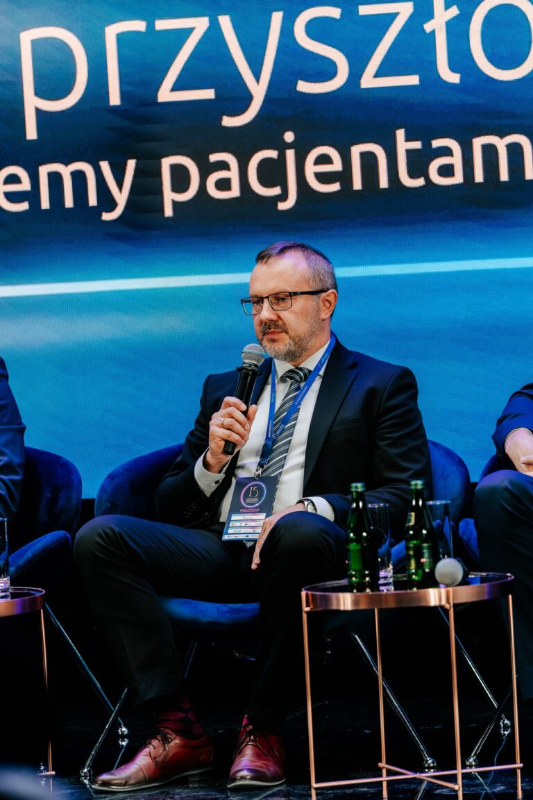 Mężczyzna w garniturze mówi do mikrofonu podczas debaty „Medycyna przyszłości: czy wszyscy będziemy pacjentami 2.0?” na 15. Forum Rozwoju Mazowsza. Siedzi na scenie na tle ekranu z tytułem dyskusji, obok stoją stoliki z butelkami wody.