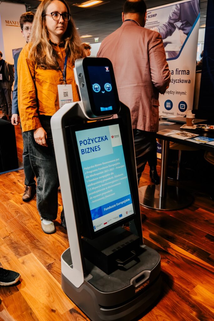 Robot z ekranem prezentujący ofertę „Pożyczka Biznes” z Funduszy Europejskich stoi w przestrzeni wystawowej podczas 15. Forum Rozwoju Mazowsza. W tle widoczni są uczestnicy wydarzenia i stoiska informacyjne.
