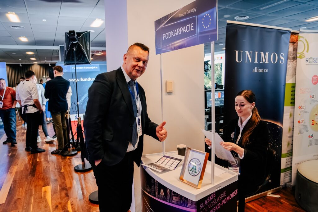 Mężczyzna w garniturze pokazuje kciuk w górę przy stoisku promującym Fundusze Europejskie dla Podkarpacia podczas 15. Forum Rozwoju Mazowsza. Za ladą siedzi kobieta z dokumentami, a w tle widoczni są inni uczestnicy wydarzenia.