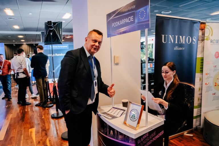 Mężczyzna w garniturze pokazuje kciuk w górę przy stoisku promującym Fundusze Europejskie dla Podkarpacia podczas 15. Forum Rozwoju Mazowsza. Za ladą siedzi kobieta z dokumentami, a w tle widoczni są inni uczestnicy wydarzenia.
