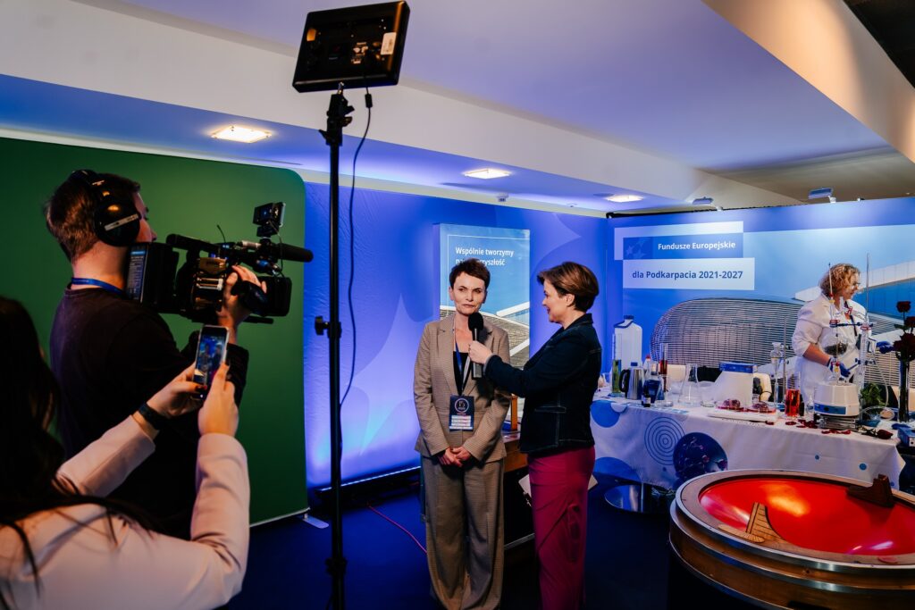 Dziennikarka przeprowadza wywiad z uczestniczką 15. Forum Rozwoju Mazowsza na tle stoiska promującego Fundusze Europejskie dla Podkarpacia 2021–2027. Obok stoi operator kamery, a w tle widoczne są eksperymenty chemiczne prowadzone przez osobę w białym fartuchu.