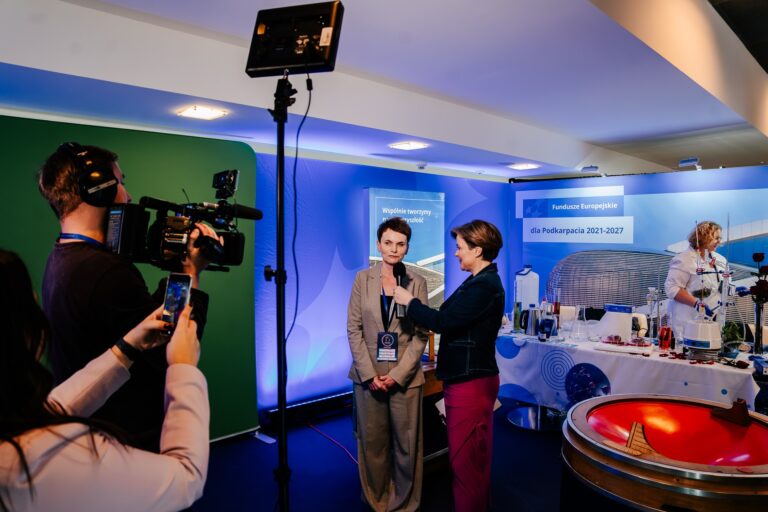 Dziennikarka przeprowadza wywiad z uczestniczką 15. Forum Rozwoju Mazowsza na tle stoiska promującego Fundusze Europejskie dla Podkarpacia 2021–2027. Obok stoi operator kamery, a w tle widoczne są eksperymenty chemiczne prowadzone przez osobę w białym fartuchu.