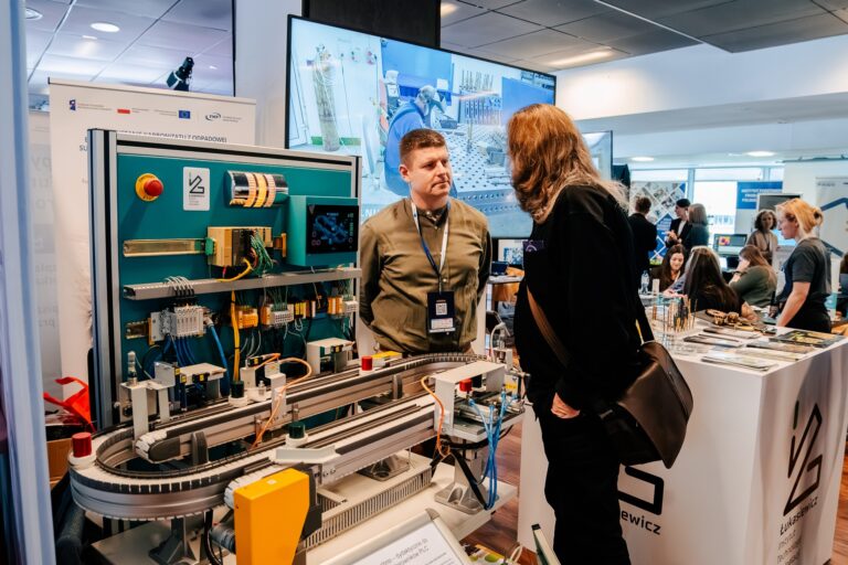 Dwie osoby rozmawiają przy stanowisku Łukasiewicz – Instytutu podczas 15. Forum Rozwoju Mazowsza. Na ekspozycji znajduje się model technologiczny z elementami automatyki i elektroniki. W tle widoczne są ekrany z wizualizacjami laboratoriów oraz inni uczestnicy wydarzenia.