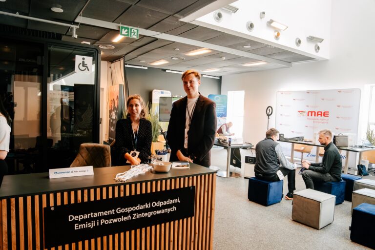 Dwie osoby stoją przy stanowisku Departamentu Gospodarki Odpadami, Emisji i Pozwoleń Zintegrowanych podczas 15. Forum Rozwoju Mazowsza. Na ladzie leżą materiały promocyjne, a w tle widać uczestników rozmawiających przy innych stoiskach.