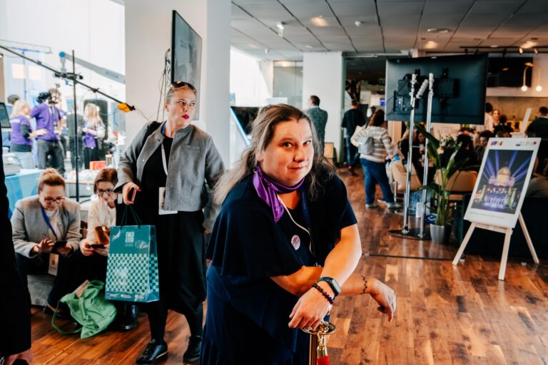 Kobieta z laską pozuje do zdjęcia w przestrzeni wydarzenia podczas 15. Forum Rozwoju Mazowsza. W tle widoczni są uczestnicy siedzący i rozmawiający przy stoiskach oraz elementy wystawowe i sprzęt nagraniowy.