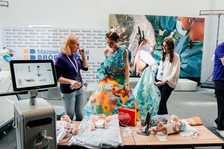 Trzy kobiety rozmawiają przy stoisku Mazowieckiego Szpitala Bródnowskiego podczas 15. Forum Rozwoju Mazowsza. Na stole leżą modele niemowląt i sprzęt medyczny, a w tle widoczne są banery oraz zdjęcie sali operacyjnej. Jedna z kobiet ma na sobie kolorową, artystyczną suknię.