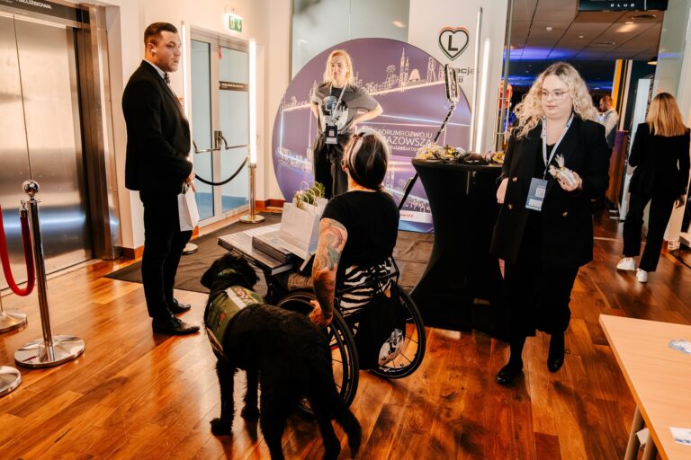 Kobieta na wózku inwalidzkim z psem asystującym rozmawia z uczestnikami wydarzenia przy stoisku 15. Forum Rozwoju Mazowsza. W tle widoczne są materiały promocyjne i baner z grafiką przedstawiającą panoramę Mazowsza.