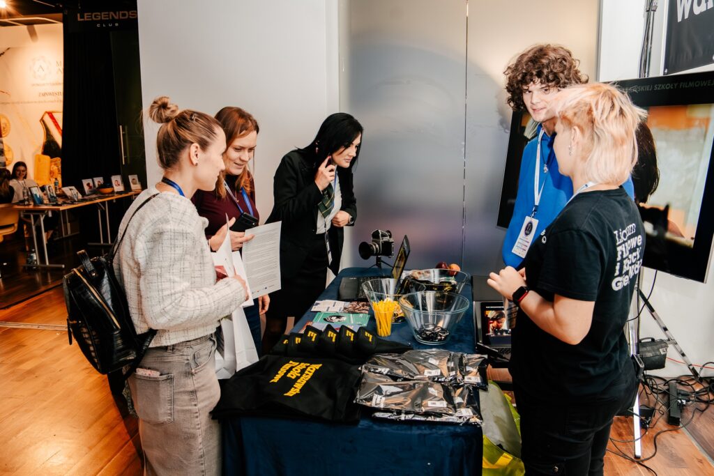 Uczestnicy rozmawiają przy stoisku Warszawskiej Szkoły Filmowej podczas 15. Forum Rozwoju Mazowsza. Na stole leżą gadżety promocyjne, ulotki i sprzęt filmowy, a w tle widać ekran i inne stoiska wydarzenia.
