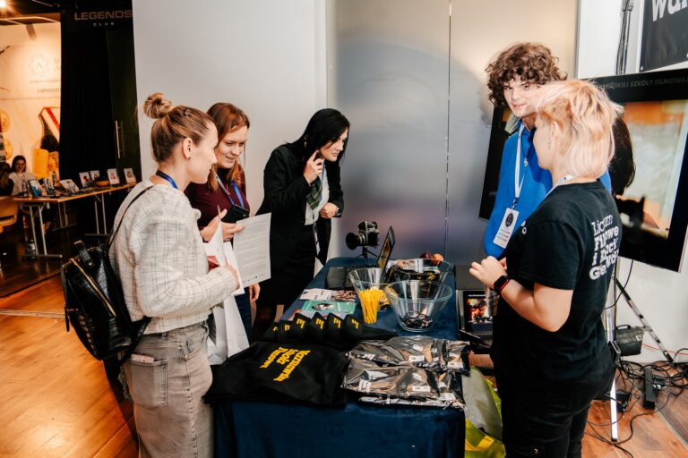Uczestnicy rozmawiają przy stoisku Warszawskiej Szkoły Filmowej podczas 15. Forum Rozwoju Mazowsza. Na stole leżą gadżety promocyjne, ulotki i sprzęt filmowy, a w tle widać ekran i inne stoiska wydarzenia.