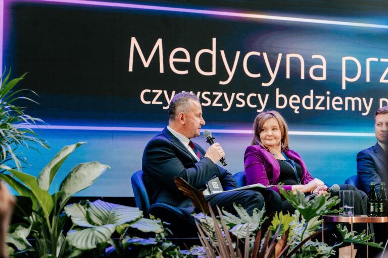 Uczestnicy debaty „Medycyna przyszłości: czy wszyscy będziemy pacjentami 2.0?” podczas 15. Forum Rozwoju Mazowsza. Mężczyzna z mikrofonem rozmawia z kobietą w fioletowej marynarce, siedzącą obok na scenie. W tle widoczny ekran z tytułem dyskusji i rośliny dekoracyjne.