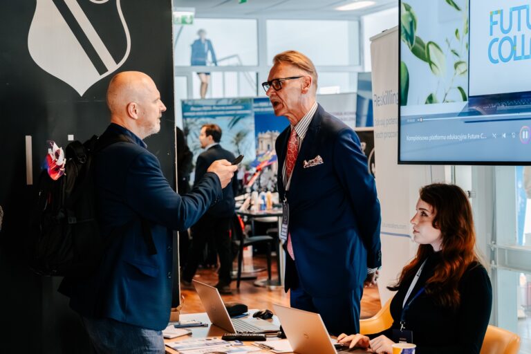 Dwaj mężczyźni rozmawiają przy stoisku Future Collars podczas 15. Forum Rozwoju Mazowsza. Jeden z nich trzyma telefon, drugi ma identyfikator na szyi. Obok nich przy biurku siedzi kobieta pracująca na laptopie. W tle widoczne są inne stoiska i uczestnicy wydarzenia.