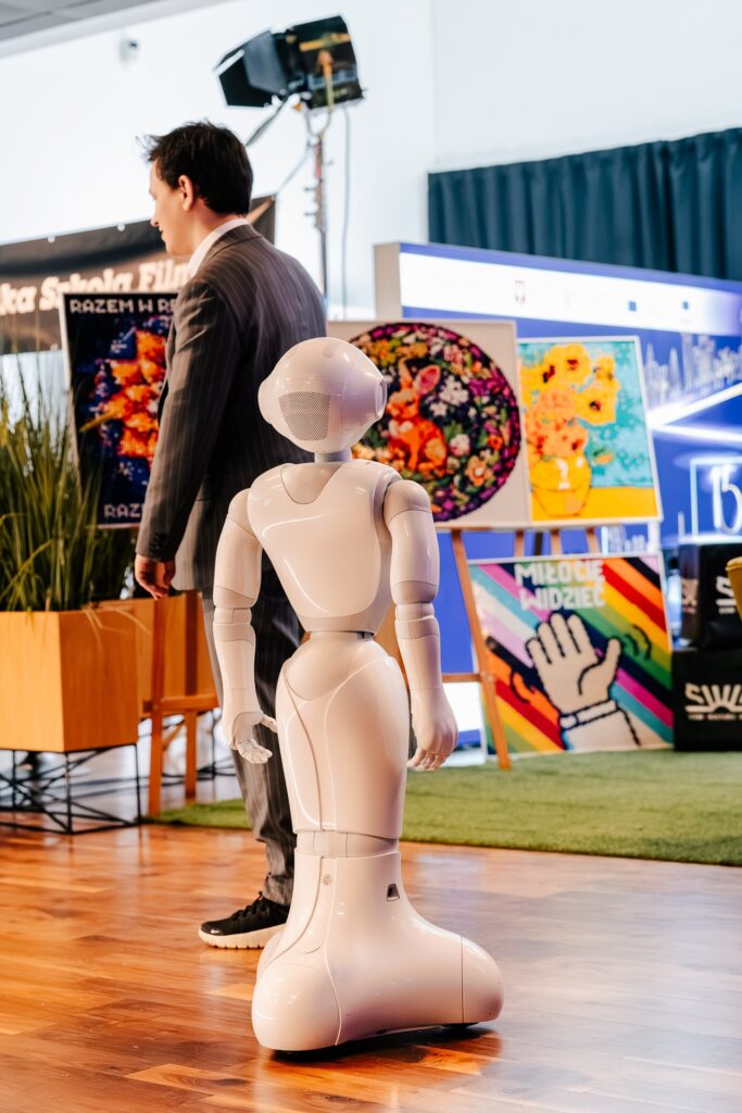 Humanoidalny robot stoi na drewnianej podłodze obok mężczyzny w garniturze podczas 15. Forum Rozwoju Mazowsza. W tle widoczne są kolorowe obrazy i plakaty wystawowe.