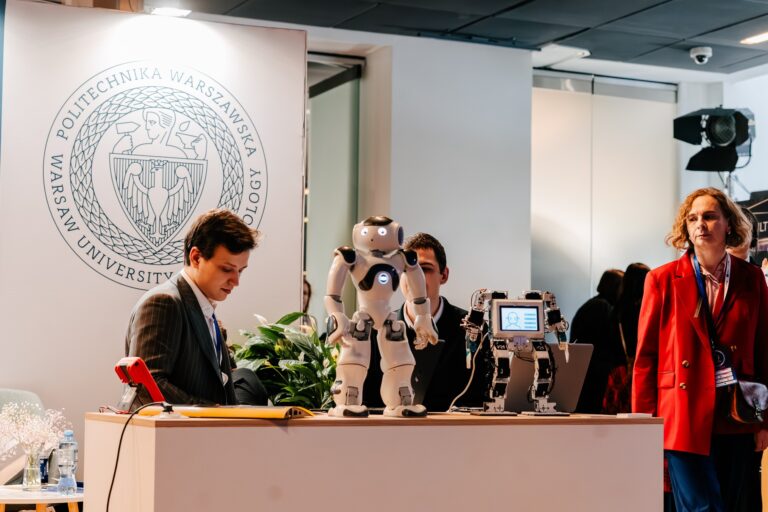 Stoisko Politechniki Warszawskiej podczas 15. Forum Rozwoju Mazowsza. Na ladzie stoją dwa humanoidalne roboty, a obok nich znajduje się dwóch mężczyzn prezentujących eksponaty. Po prawej stronie stoi kobieta w czerwonej marynarce.