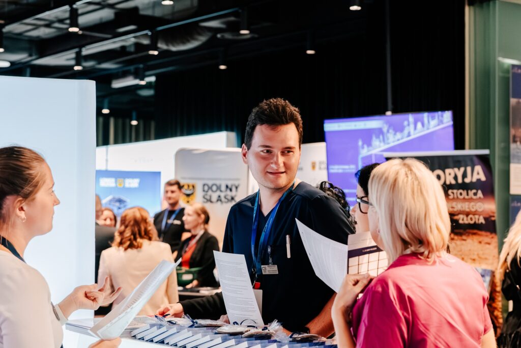 Grupa osób rozmawia przy stoisku wystawowym podczas 15. Forum Rozwoju Mazowsza. Mężczyzna z identyfikatorem trzyma dokumenty i prowadzi rozmowę z dwiema kobietami. W tle widoczne są inne stoiska i uczestnicy wydarzenia.