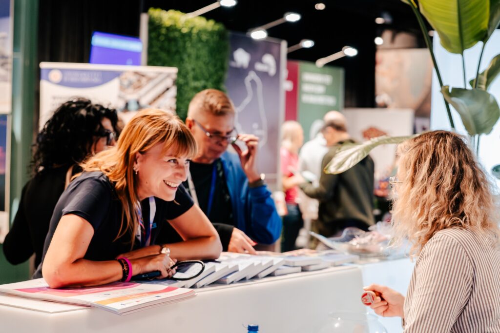 Uśmiechnięta kobieta rozmawia z uczestniczką przy stoisku wystawowym podczas 15. Forum Rozwoju Mazowsza. W tle widać inne osoby i kolorowe ścianki informacyjne różnych instytucji.