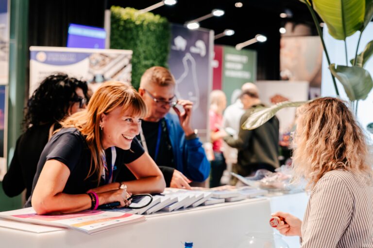 Uśmiechnięta kobieta rozmawia z uczestniczką przy stoisku wystawowym podczas 15. Forum Rozwoju Mazowsza. W tle widać inne osoby i kolorowe ścianki informacyjne różnych instytucji.
