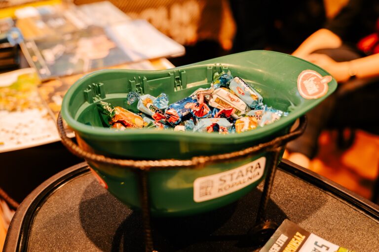 Zielony kask z logo „Stara Kopalnia” wypełniony kolorowymi cukierkami, stojący na stole podczas 15. Forum Rozwoju Mazowsza. W tle widoczne są ulotki i materiały promocyjne.